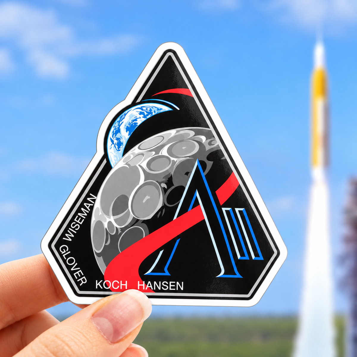 Artemis II mission badge vinyl sticker held in hand in front of a rocket launch